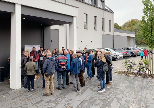 Eine größere Gruppe Menschen versammelt sich vor einem Gebäude, im Hintergrund steht ein Tafel zum Stadtteilspaziergang.